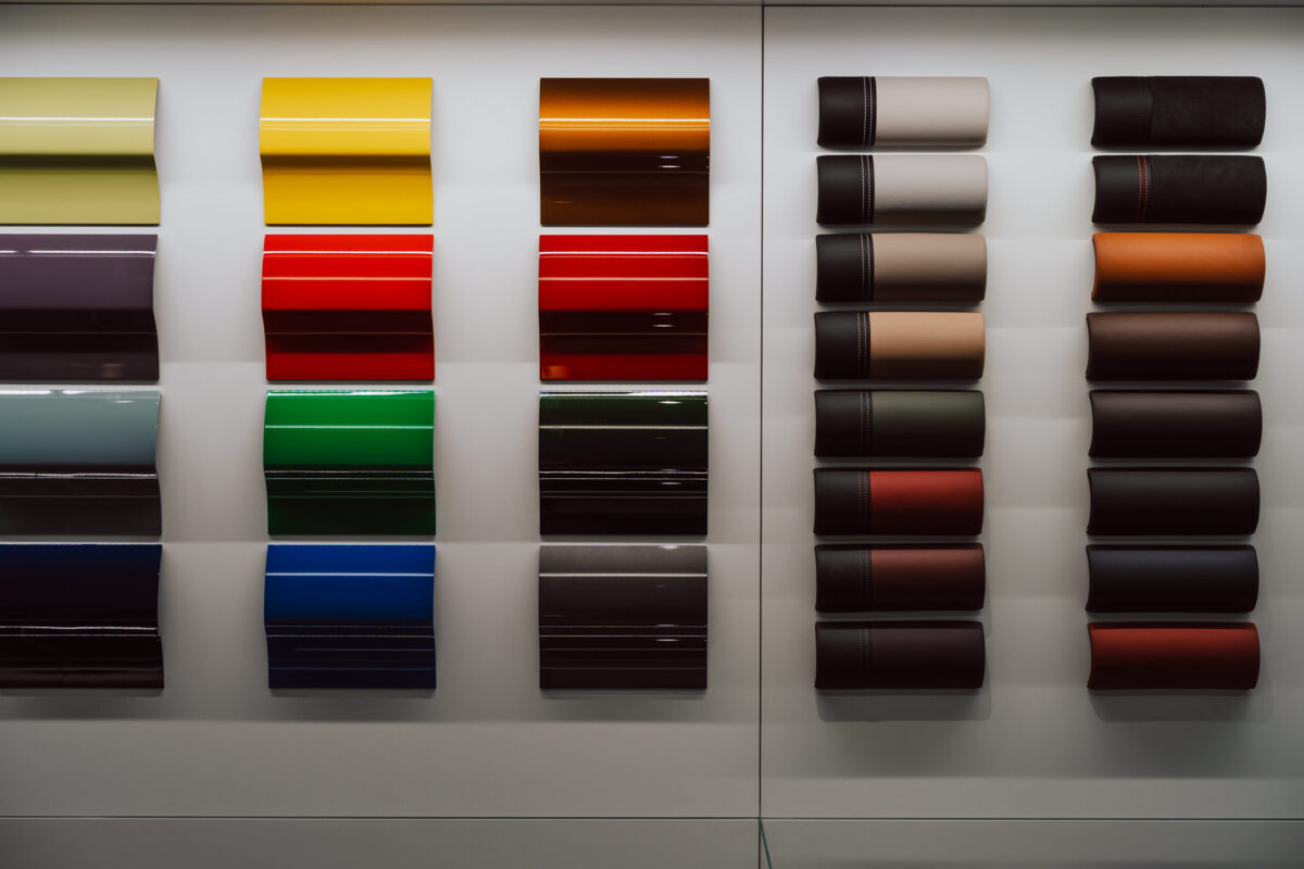 Auf der linken Seite sind rechteckige Muster von glänzenden Autolacken in verschiedenen Farben ausgestellt, während auf der rechten Seite Rollen mit verschiedenfarbigen Materialien für die Autoinnenausstattung angeordnet sind - ein elegantes Schaufenster für die Eröffnung des Showrooms, alles an einer weißen Wand.