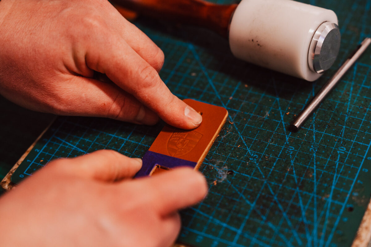 Eine Person drückt mit ihren Fingern auf einer grünen Schneidematte ein Design in ein Stück Leder, während ein weißer Hammer und ein Metallwerkzeug in der Nähe sind - vielleicht ein individuelles Schild für eine Saloneröffnung.