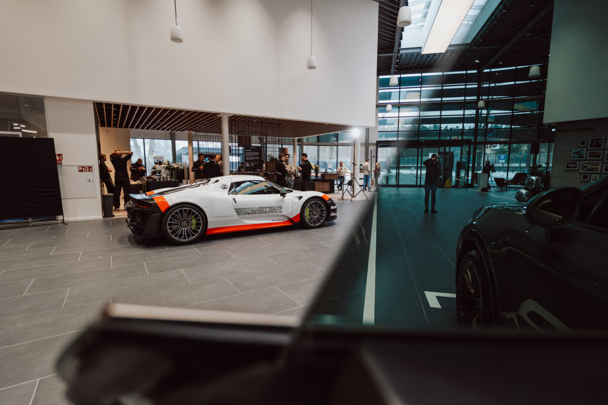 Der weiß-orangefarbene Sportwagen wird in einem modernen Showroom ausgestellt, mit großen Fenstern, grauen Bodenfliesen und ein paar Menschen im Hintergrund, von denen einige Fotos machen.