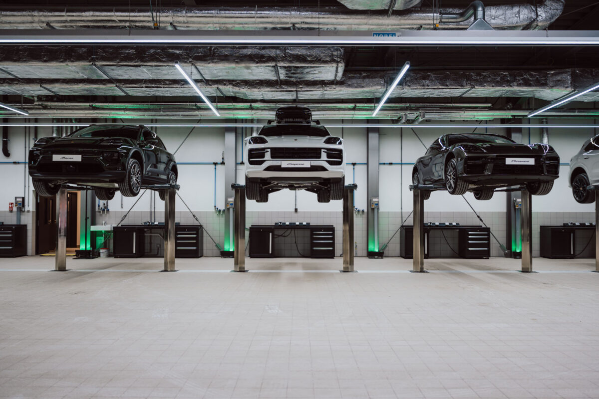 Vier Autos, darunter zwei schwarze und ein weißes, stehen auf Wagenhebern in einer modernen, sauberen Autowerkstatt mit Werkzeugschränken und freiliegenden Deckenschächten - bereit für die Eröffnung des Ausstellungsraums. Die grüne Akzentbeleuchtung hebt den Raum im Hintergrund hervor. 