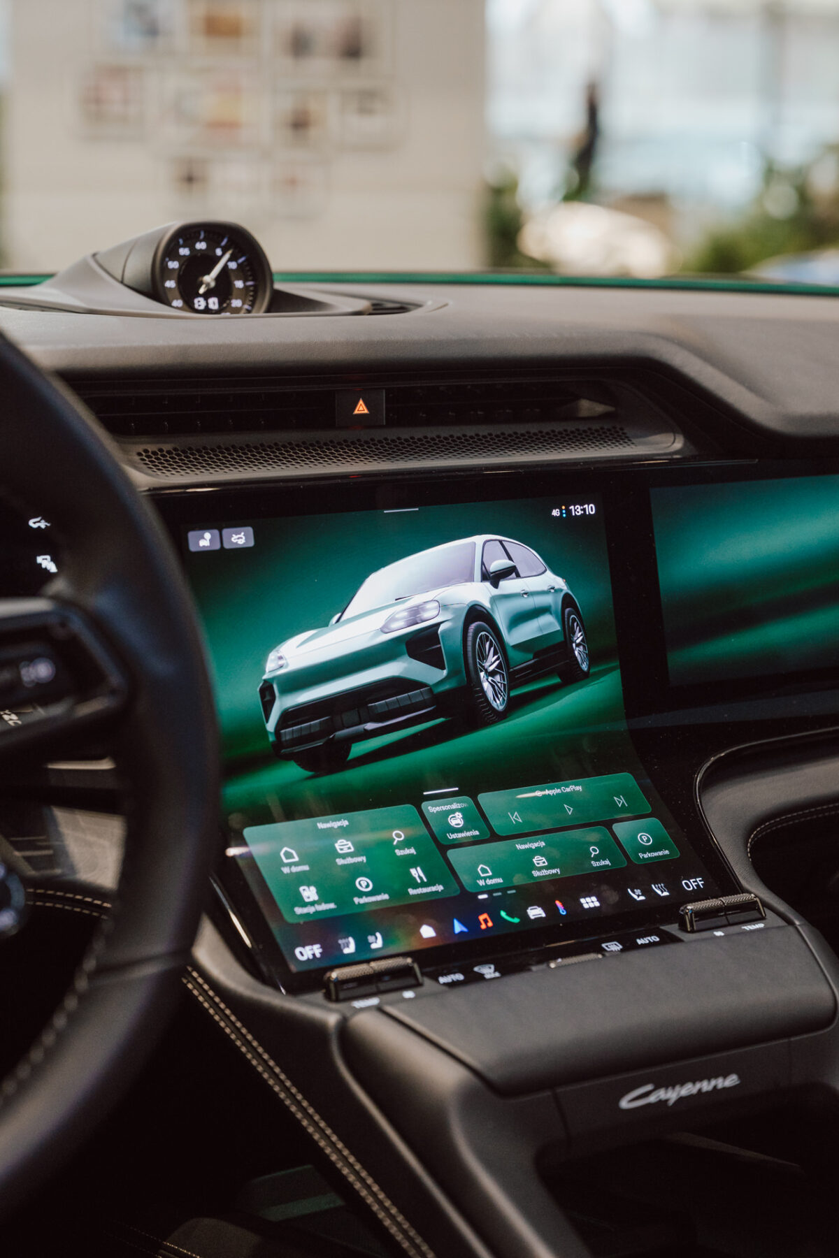 Nahaufnahme des digitalen Touchscreen-Armaturenbretts des Fahrzeugs mit einem Bild des grünen SUV, verschiedenen Bediensymbolen und einem Teil des Lenkrads und der Uhr. Auf der Konsole steht das Wort "Cayenne", das bei der Eröffnung des Ausstellungsraums aufgenommen wurde. 