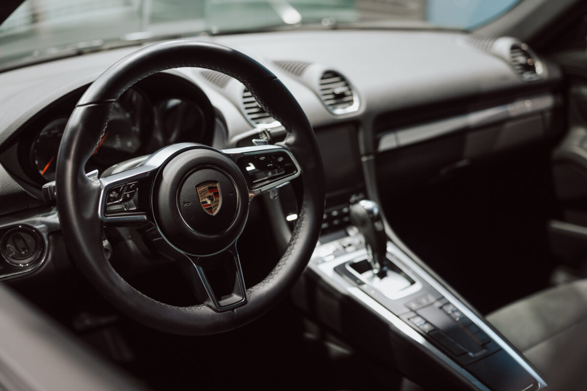 Nahaufnahme des Interieurs des Luxuswagens im offenen Showroom, mit einem schwarzen Lederlenkrad mit Porsche-Logo, einem modernen Armaturenbrett, einer Mittelkonsole und eleganten schwarzen Sitzen.