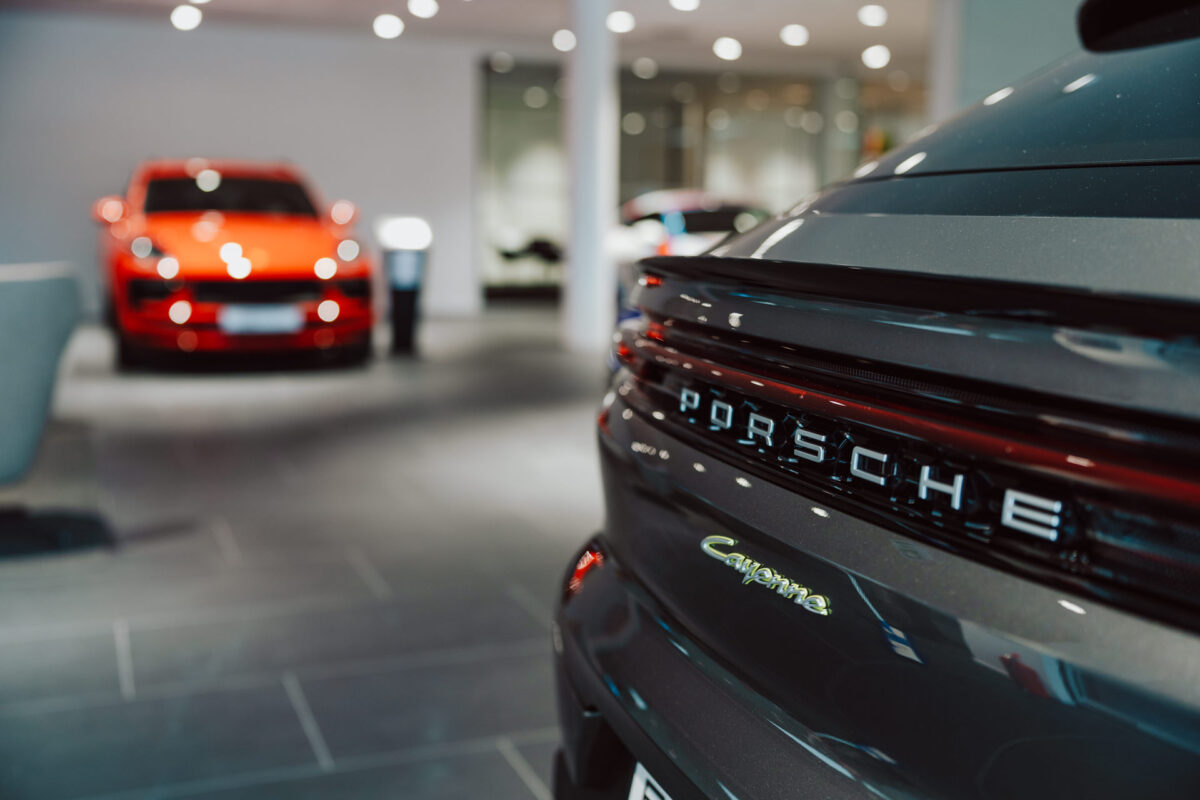 Nahaufnahme einer schwarzen Porsche Cayenne-Plakette in einem modernen Autohaus während der Eröffnung des Showrooms, wobei das rote Auto im Hintergrund bei heller Beleuchtung verschwimmt.