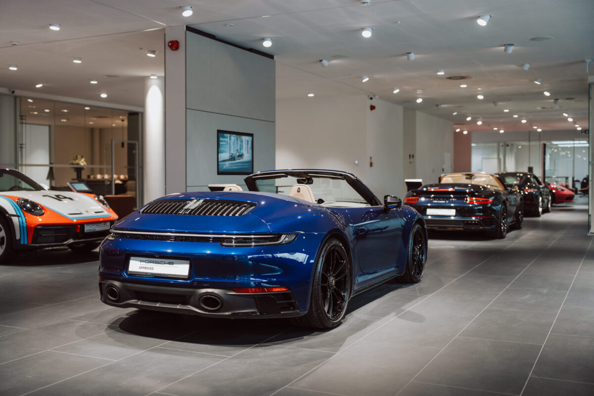 Das blaue Porsche Sportcabrio wird bei der Eröffnung des Showrooms in einem eleganten, modernen Ausstellungsraum mit heller Beleuchtung und einem sauberen, minimalistischen Design neben anderen Luxuswagen präsentiert.