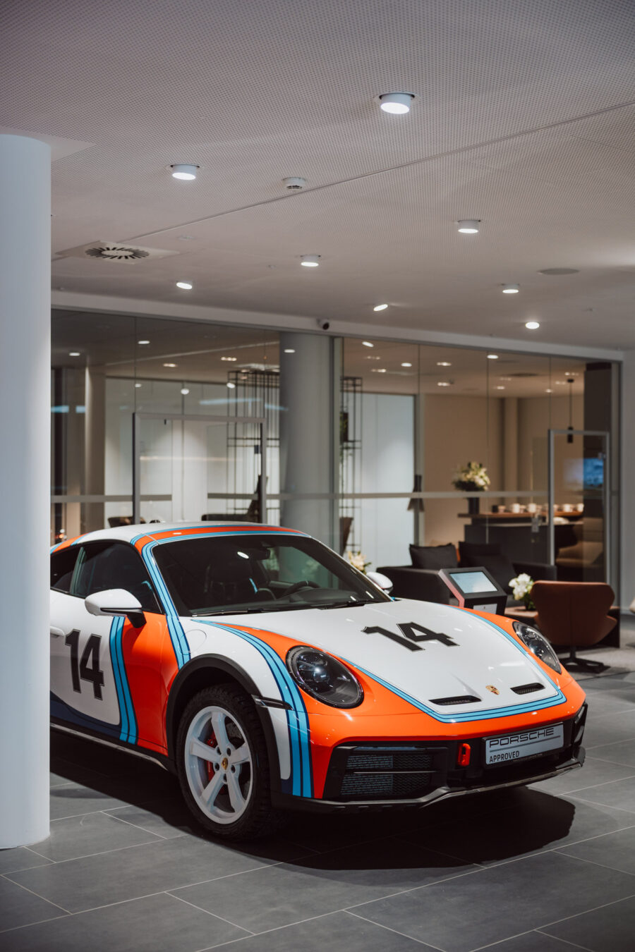 Ein weißer Porsche Sportwagen mit roten, blauen und schwarzen Rennstreifen und der Nummer 14 auf der Motorhaube und an der Seite, präsentiert in einer offenen Limousine in einem modernen, gut beleuchteten Ausstellungsraum.