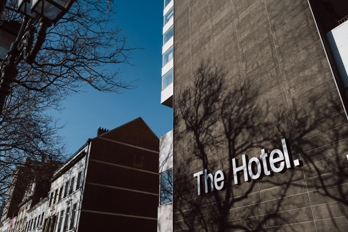 Wysoki budynek z napisem "The Hotel" na ścianie, rzucający ostre cienie drzew. Bezlistne gałęzie rozciągają się na czystym, błękitnym niebie, ze starszymi budynkami widocznymi po lewej stronie.