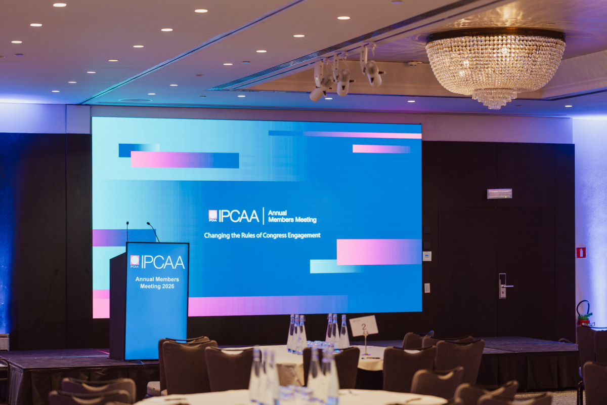 Nowoczesna sala konferencyjna z okrągłymi stołami, butelkami wody na stołach i dużym ekranem wyświetlającym "IPCAA Annual Members Meeting: Zmiana zasad zaangażowania Kongresu".