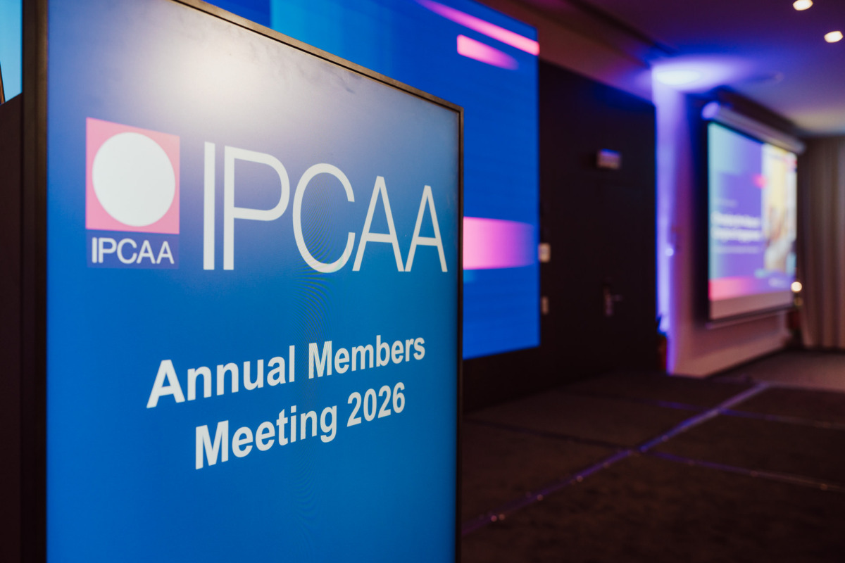Podium z napisem "IPCAA Annual Members Meeting 2026" w sali konferencyjnej z niebieskim i fioletowym oświetleniem oraz ekranem prezentacyjnym widocznym w tle.