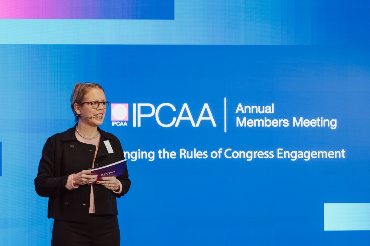Kobieta stoi na scenie trzymając teczkę, przemawiając na dorocznym spotkaniu członków IPCAA. W tle wyświetlany jest tytuł wydarzenia i slogan "Changing the Rules of Congress Engagement" na niebieskim ekranie.
