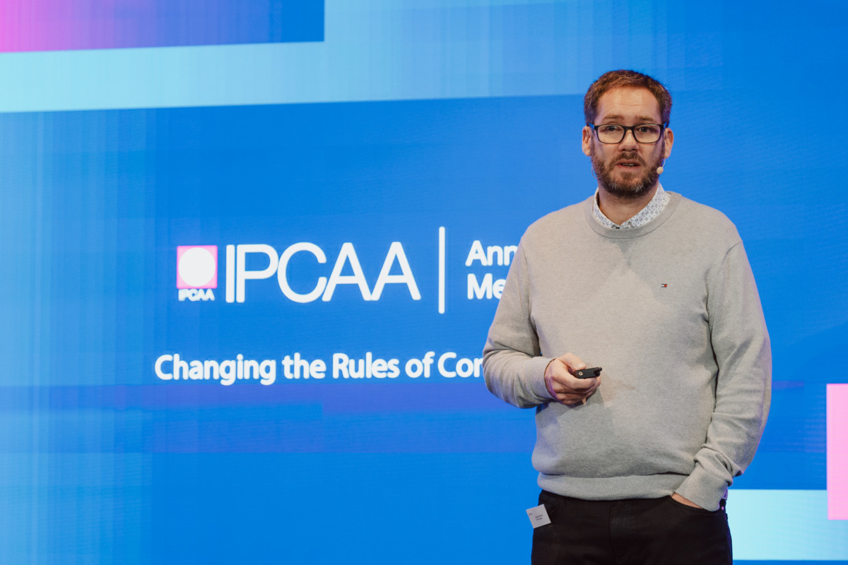 Mężczyzna w okularach i jasnoszarym swetrze przemawia na scenie trzymając pilota, na niebieskim tle wyświetlany jest tekst "IPCAA Annual Meeting: Zmiana zasad kongresów".