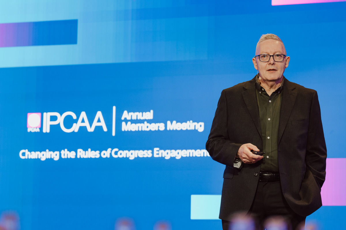 Osoba w ciemnym garniturze stoi na scenie, trzymając pilota, a za nią znajduje się ekran z napisem "IPCAA Annual Members Meeting-Changing the Rules of Congress Engagement".