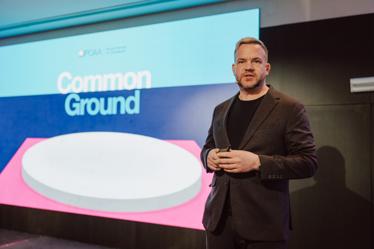 Mężczyzna w ciemnym garniturze stoi na scenie, trzymając urządzenie, z dużym ekranem za nim wyświetlającym słowa "Common Ground" w białym tekście na niebieskim i różowym tle.