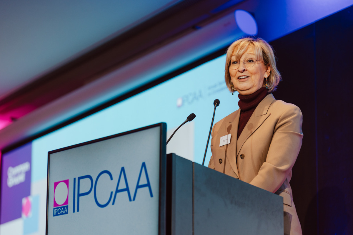 Kobieta w brązowej marynarce i okularach przemawia na podium z napisem "IPCAA" w otoczeniu konferencyjnym, z ekranem projekcyjnym w tle wyświetlającym to samo logo.