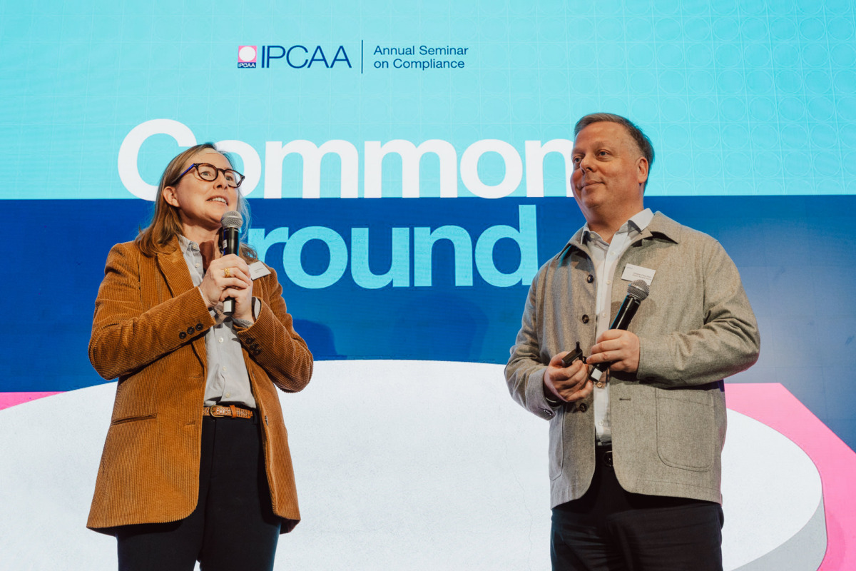 Dwie osoby z mikrofonami stoją na scenie podczas dorocznego seminarium IPCAA na temat zgodności, przemawiając przed ekranem z napisem "Common Ground". Obie osoby ubrane są w stroje biznesowe.