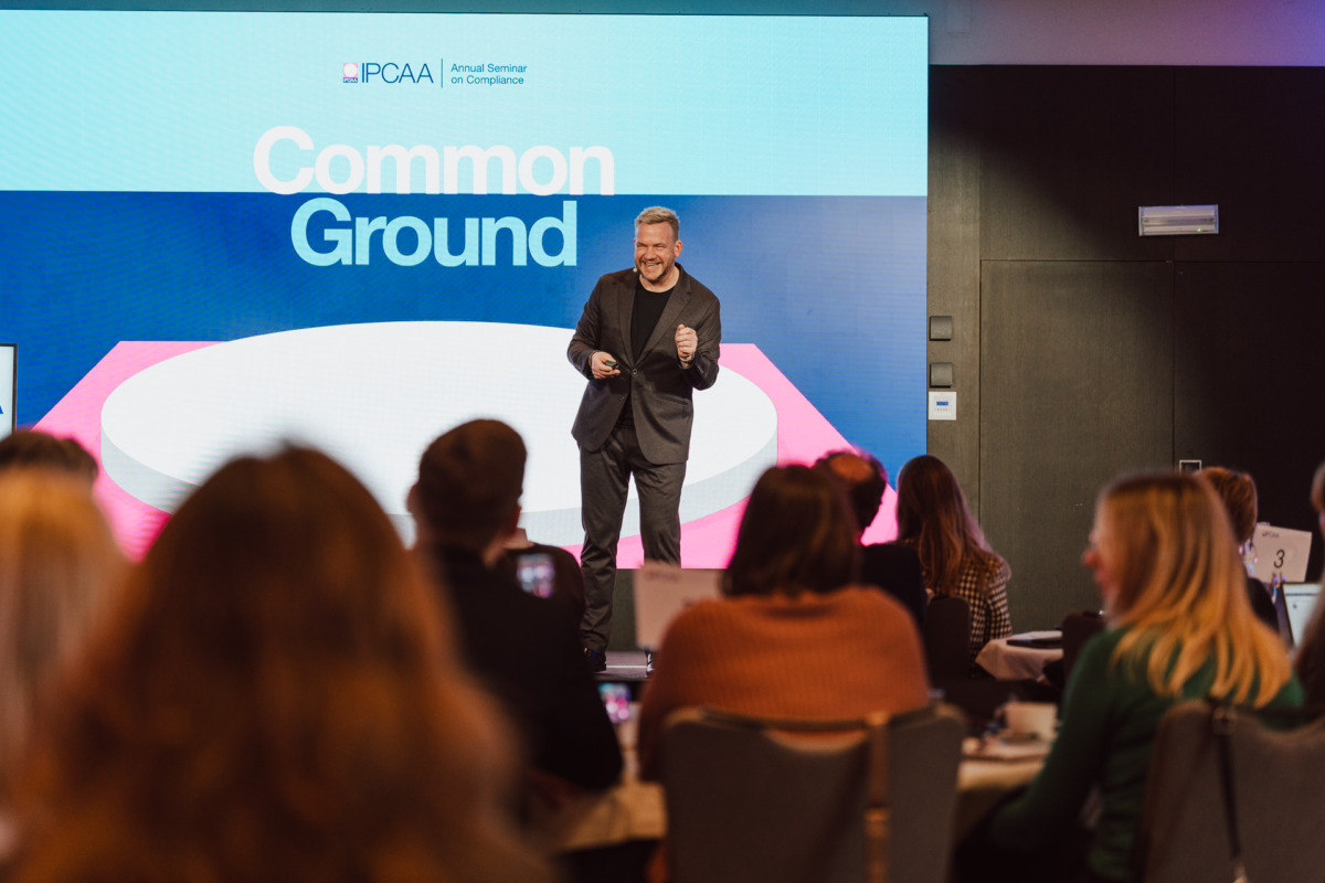 Prelegent stoi na scenie przed ekranem wyświetlającym "Common Ground" podczas seminarium, podczas gdy publiczność siedzi przy okrągłych stołach i uważnie słucha.