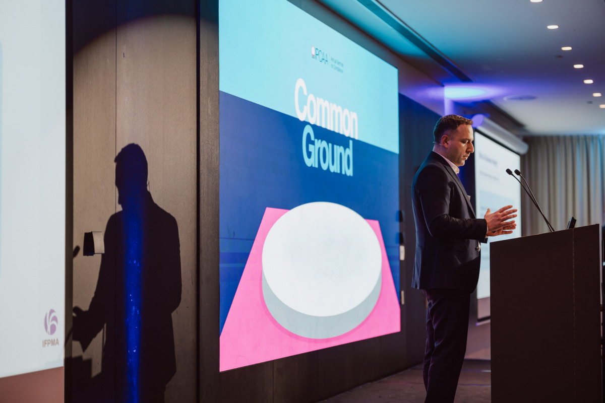 Mężczyzna w garniturze stoi na podium, przemawiając na konferencji. Za nim znajduje się duży ekran wyświetlający słowa "Common Ground" z kolorową grafiką. Pomieszczenie jest jasno oświetlone reflektorami.
