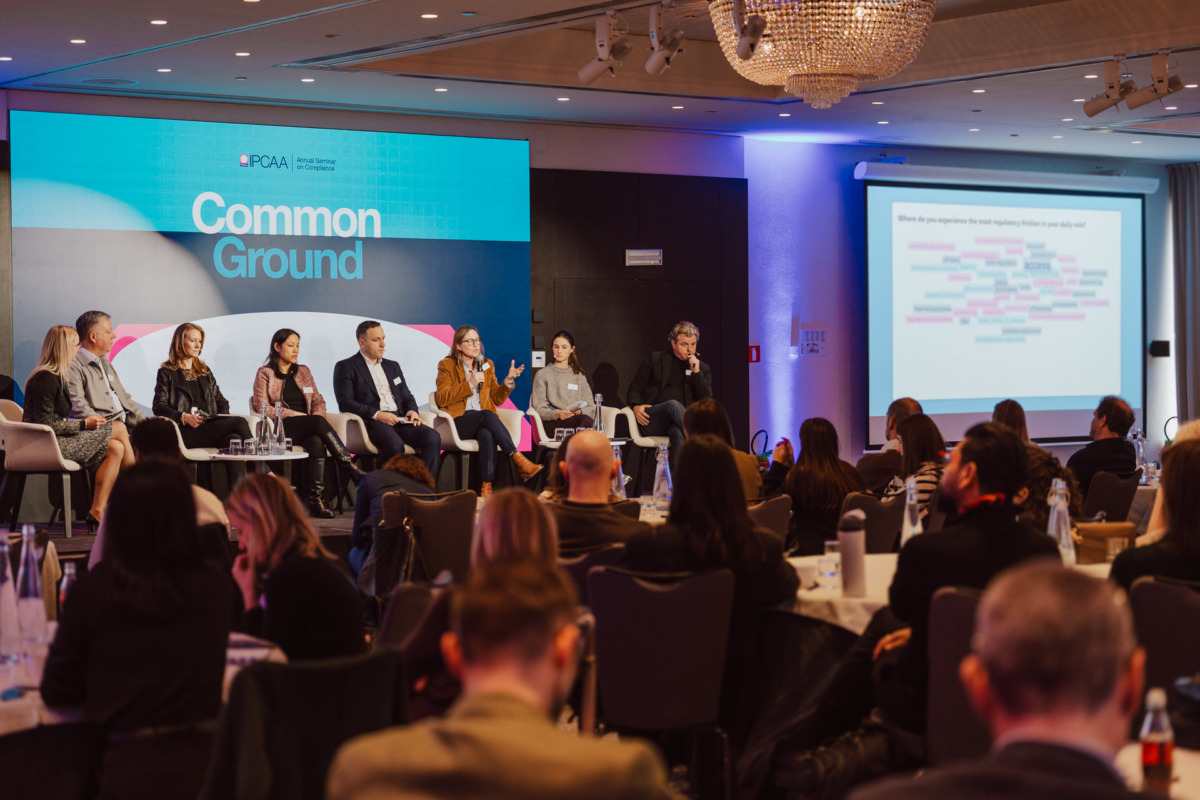 Panel ośmiu mówców siedzi na scenie przed dużym ekranem wyświetlającym "Common Ground", zwracając się do publiczności w nowoczesnej sali konferencyjnej z żyrandolem nad głową.