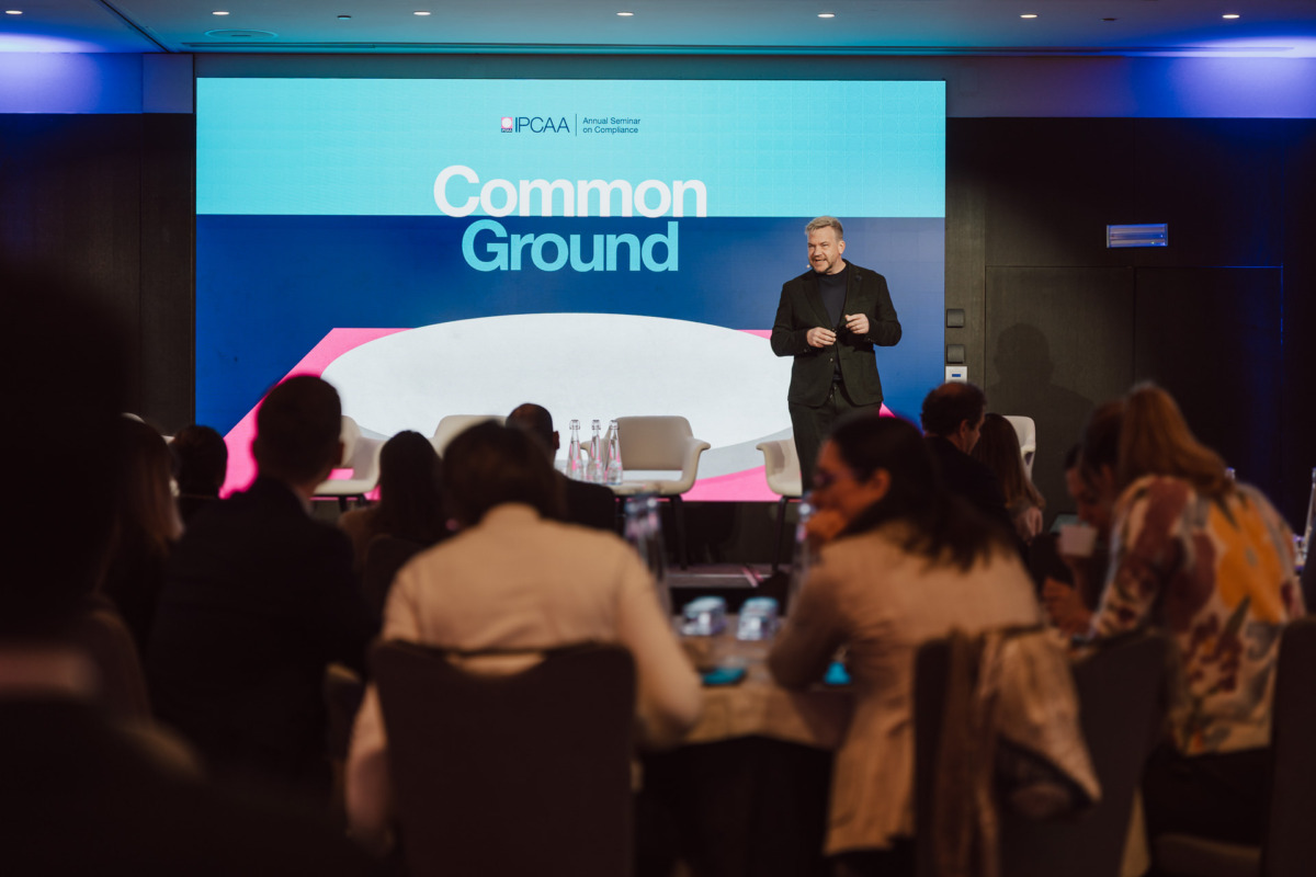 Prelegent stoi na scenie przed dużym ekranem wyświetlającym "Common Ground" na konferencji, zwracając się do publiczności siedzącej przy okrągłych stołach.