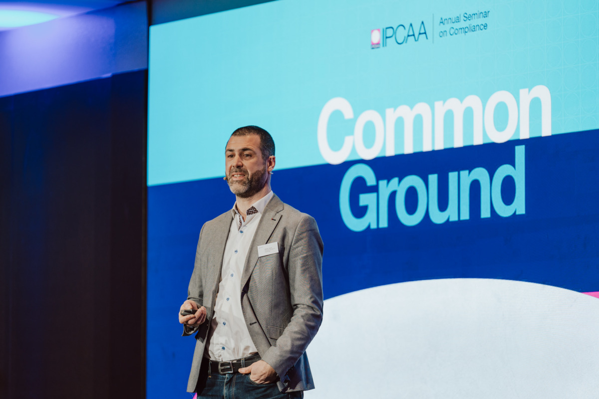 Mężczyzna w szarej marynarce przemawia na scenie podczas seminarium, stojąc przed dużym ekranem z napisem "IPCAA Annual Seminar on Compliance" i "Common Ground.