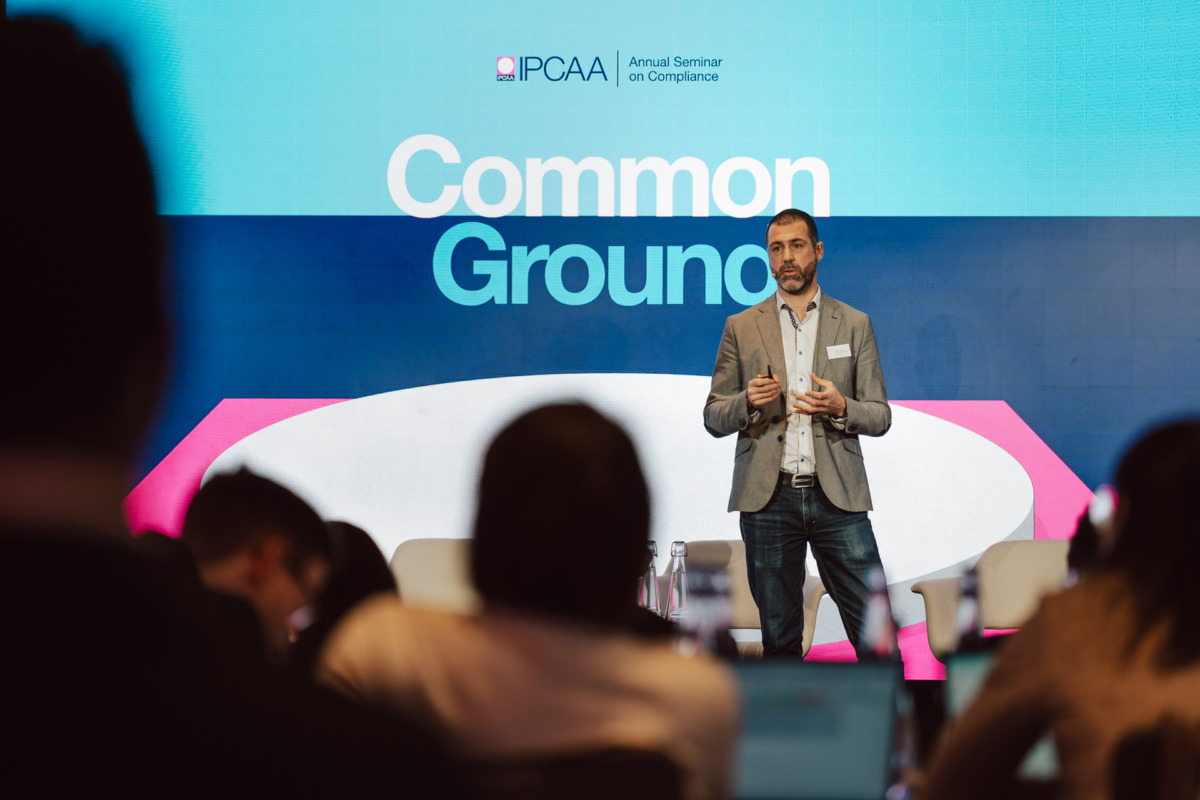 Mężczyzna w marynarce stoi na scenie przemawiając podczas seminarium zatytułowanego "Common Ground", a za nim znajduje się baner IPCAA Annual Seminar on Compliance. Członkowie publiczności są widoczni na pierwszym planie.