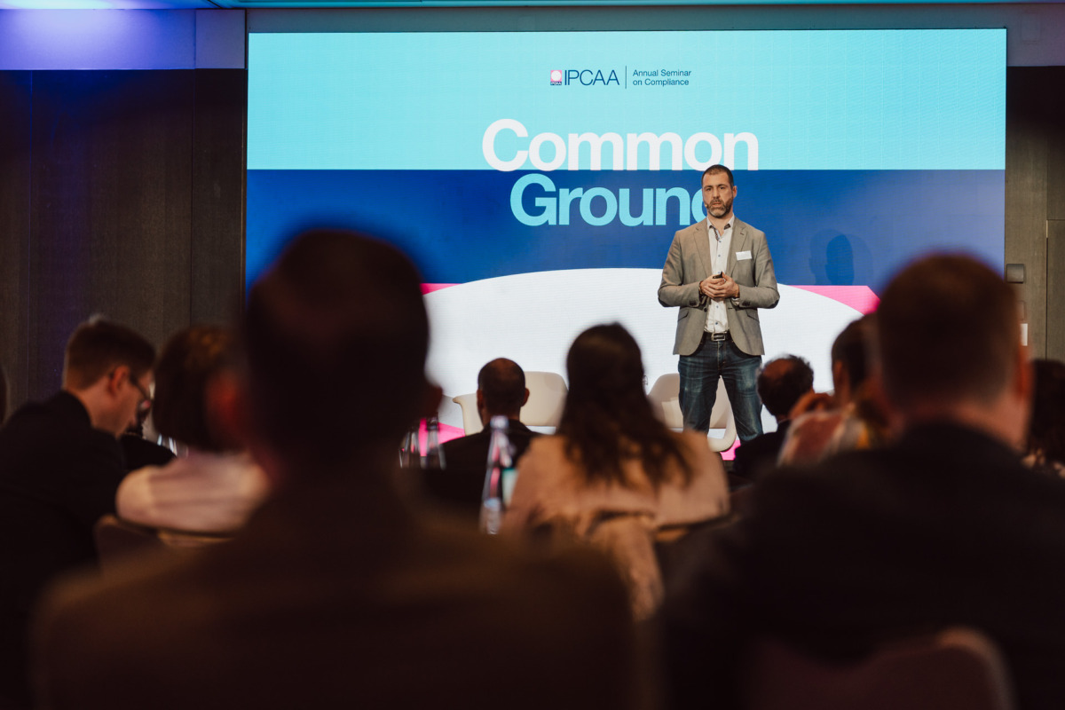 Mężczyzna w szarej marynarce stoi na scenie i przemawia do publiczności. Za nim na dużym ekranie wyświetlany jest napis "Common Ground" i "PCAA Annual Seminar on Compliance".