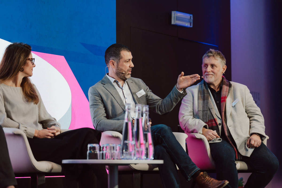 Trzy osoby siedzą na scenie podczas panelu dyskusyjnego; mężczyzna w centrum gestykuluje podczas mówienia, podczas gdy kobieta po jego lewej i mężczyzna po jego prawej stronie słuchają. Na pierwszym planie znajduje się stół ze szklankami wody.