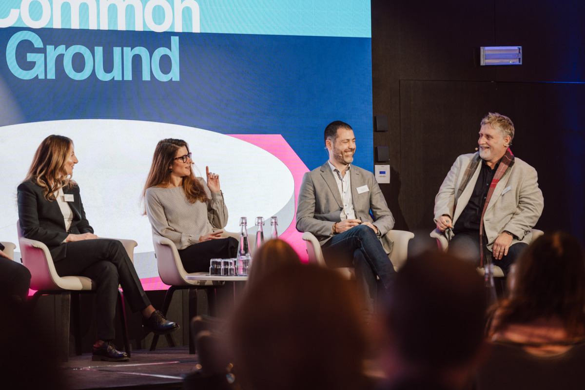 Cztery osoby siedzą na scenie na krzesłach, uśmiechając się i rozmawiając podczas panelu dyskusyjnego. Duży ekran za nimi wyświetla słowa "Common Ground". Butelki z wodą stoją na małym stoliku przed nimi.