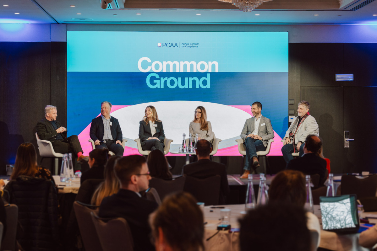 Pięciu panelistów siedzących na scenie przed ekranem z napisem "Common Ground", przemawiających na konferencji. Członkowie publiczności siedzą przy stolikach na pierwszym planie, słuchając i robiąc notatki.