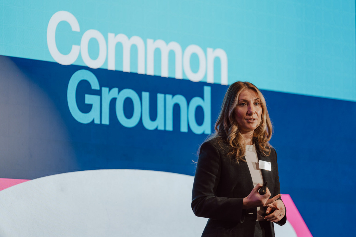 Kobieta w garniturze przemawia na scenie przed dużym ekranem, na którym pogrubioną czcionką wyświetlany jest napis "Common Ground".