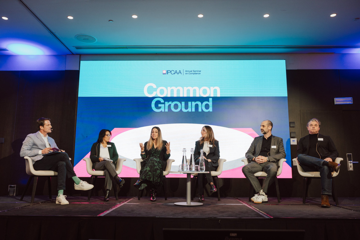 Sześć osób siedzi na scenie na nowoczesnych krzesłach, prowadząc dyskusję panelową przed dużym ekranem wyświetlającym słowa "Common Ground" i logo PCAA. Wyglądają na zaangażowanych, a jeden z mówców gestykuluje podczas rozmowy.