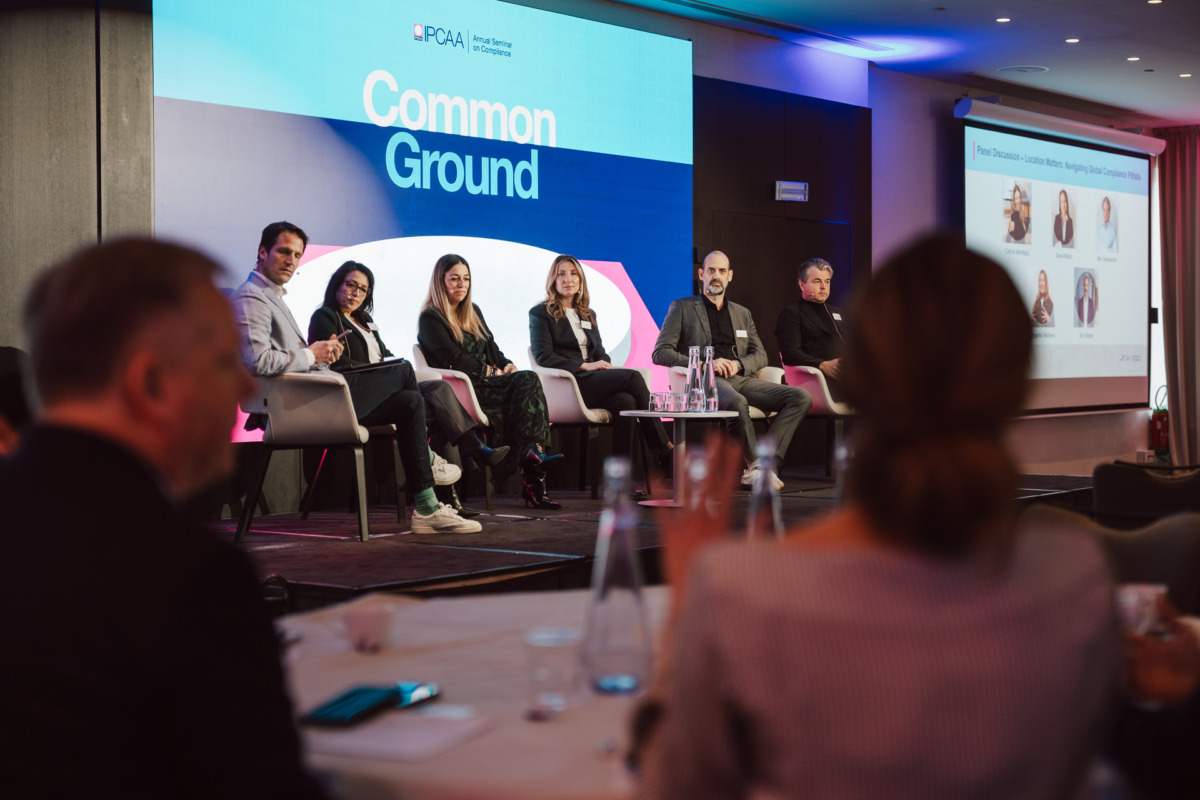 Panel sześciu osób siedzi na scenie przed ekranem z napisem "Common Ground", przemawiając do publiczności na konferencji. Pokój ma fioletowe oświetlenie, a uczestnicy siedzą przy stołach na pierwszym planie.