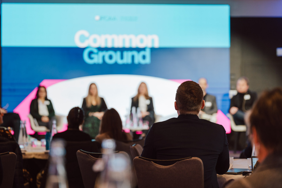 Grupa ludzi siedzi na panelu na scenie w celu przeprowadzenia dyskusji, a za nimi na dużym ekranie wyświetlany jest komunikat "Common Ground". Publiczność siedzi na pierwszym planie, twarzą do sceny.