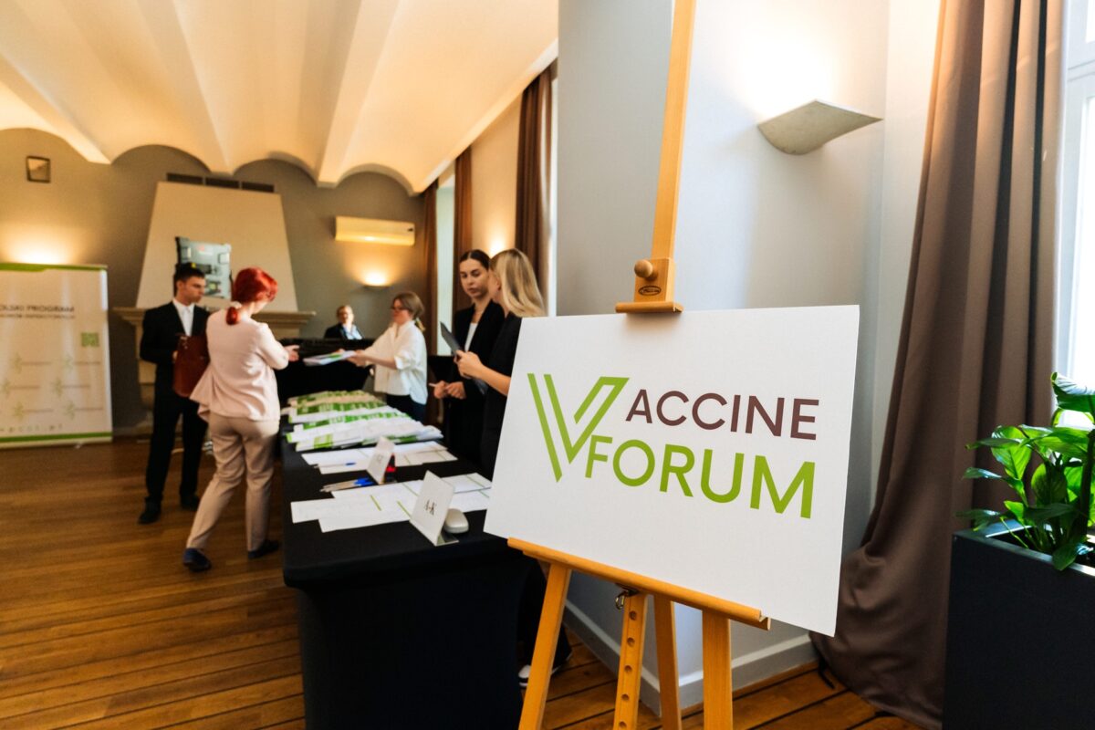 Tablica z logo Vaccine Forum na sztaludze, w tle rejestracja gości konferencji w Villi Foksal