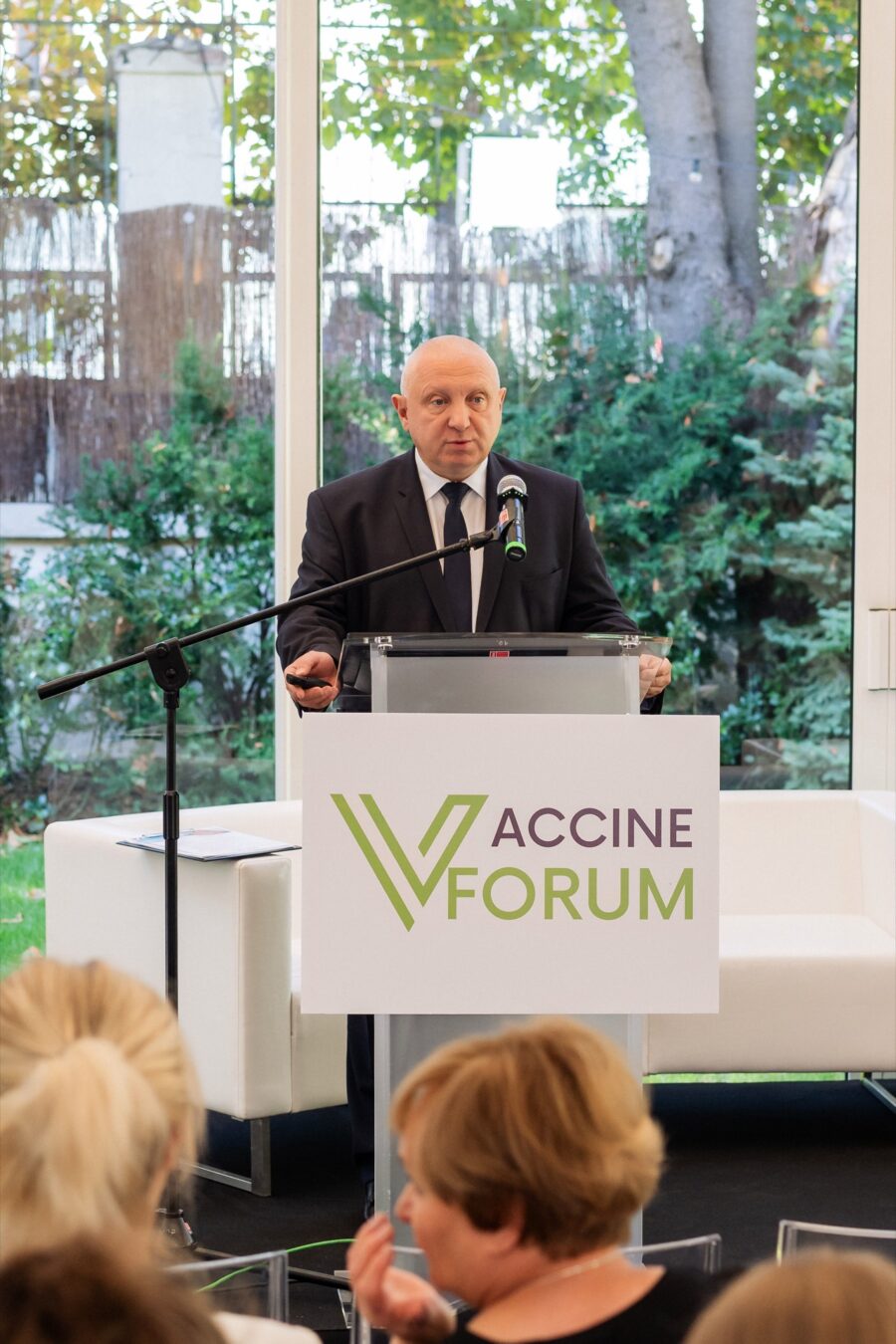 Prelegent w granatowym garniturze przemawia przy mównicy z logo Vaccine Forum