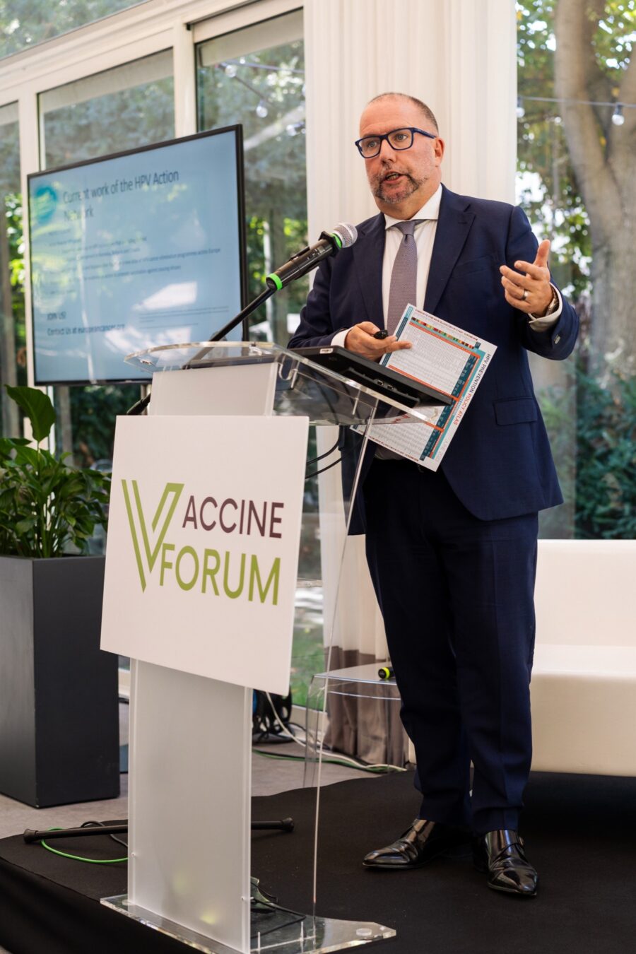 Ekspert z broszurą w dłoni przemawia przy mównicy z logo Vaccine Forum