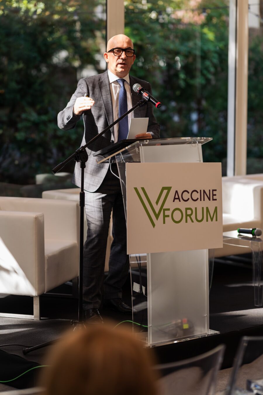Prelegent w szarym garniturze w kratę gestykuluje przy mównicy Vaccine Forum